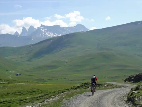 Cyclist riding along a gravel track on the Plateau d'Emparis with the peaks of the Écrins massif in the background.