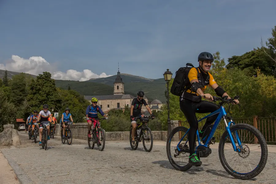 Grupo de ciclistas cruzando el Puente del Perdón, en Rascafría, con el Monasterio de El Paular y las montañas del Valle del Lozoya al fondo